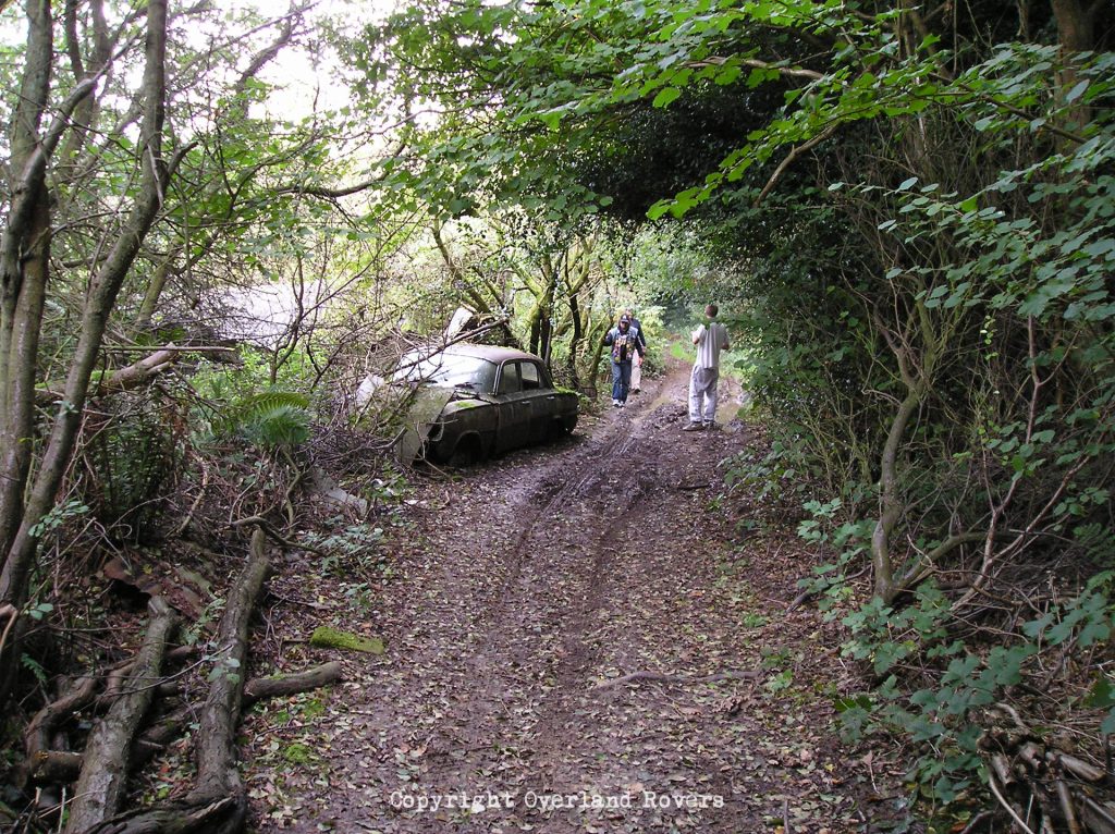 Looking down a dirt track with undergrowth either side. To the left is a decaying Vauxhall Victor classic car that's been abandoned