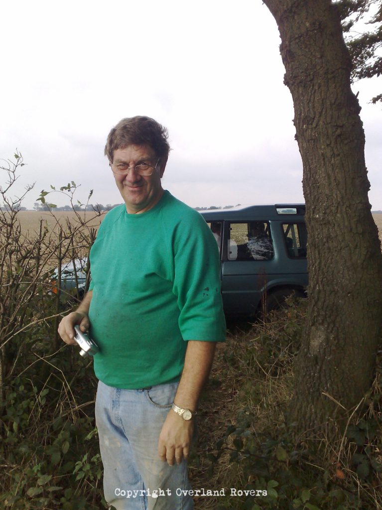 A man in a green top, jeans, and glasses, peering at the camera, with a tree to his left and a blue Land Rover Discovery 1 in the background