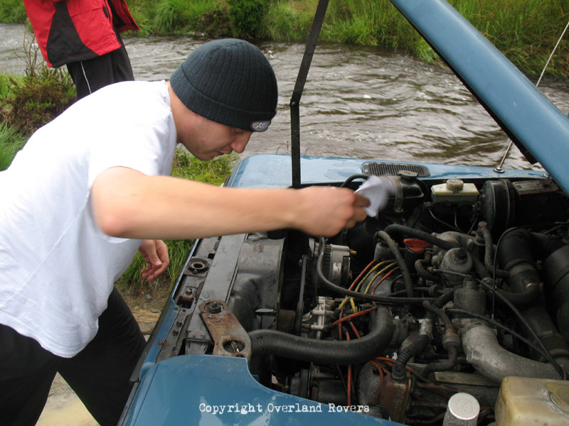 A person under the raised bonnet of a blue Land Rover 110 CSW. A fast flowing stream is in the background