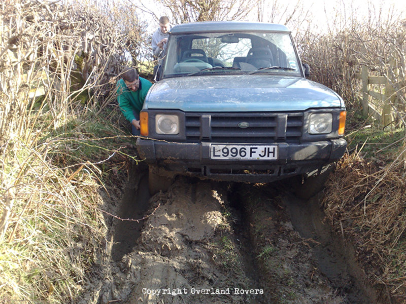 A blue Land Rover Discovery 1, stuck up to its axles in mud. There are bushes tight on both sides and a couple of men trying to recover the vehicle