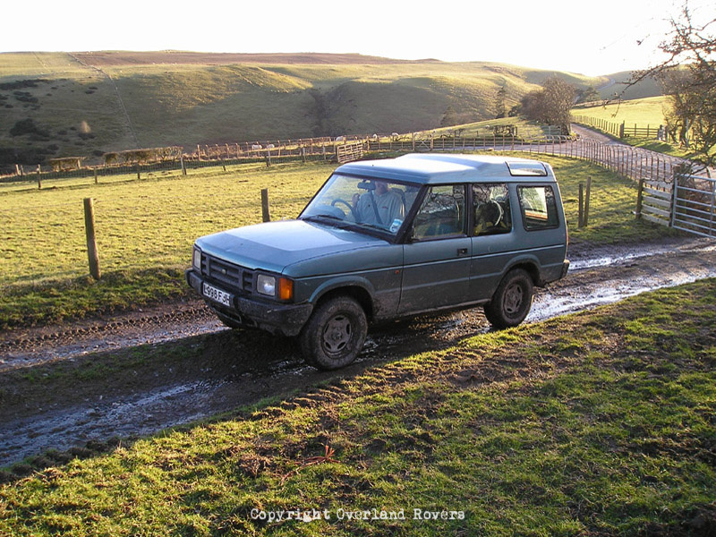 A blue Land Rover Discovery 1, 3 door, set on a gravel track with rolling green hills behind it.
