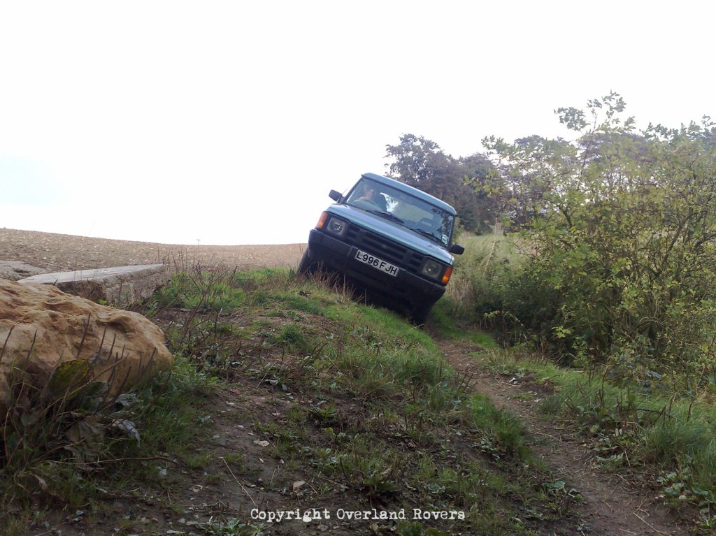 A blue Land Rover Discovery 1, on a dirt trail, with bushes to the right. The vehicle is leaning heavily to their left.