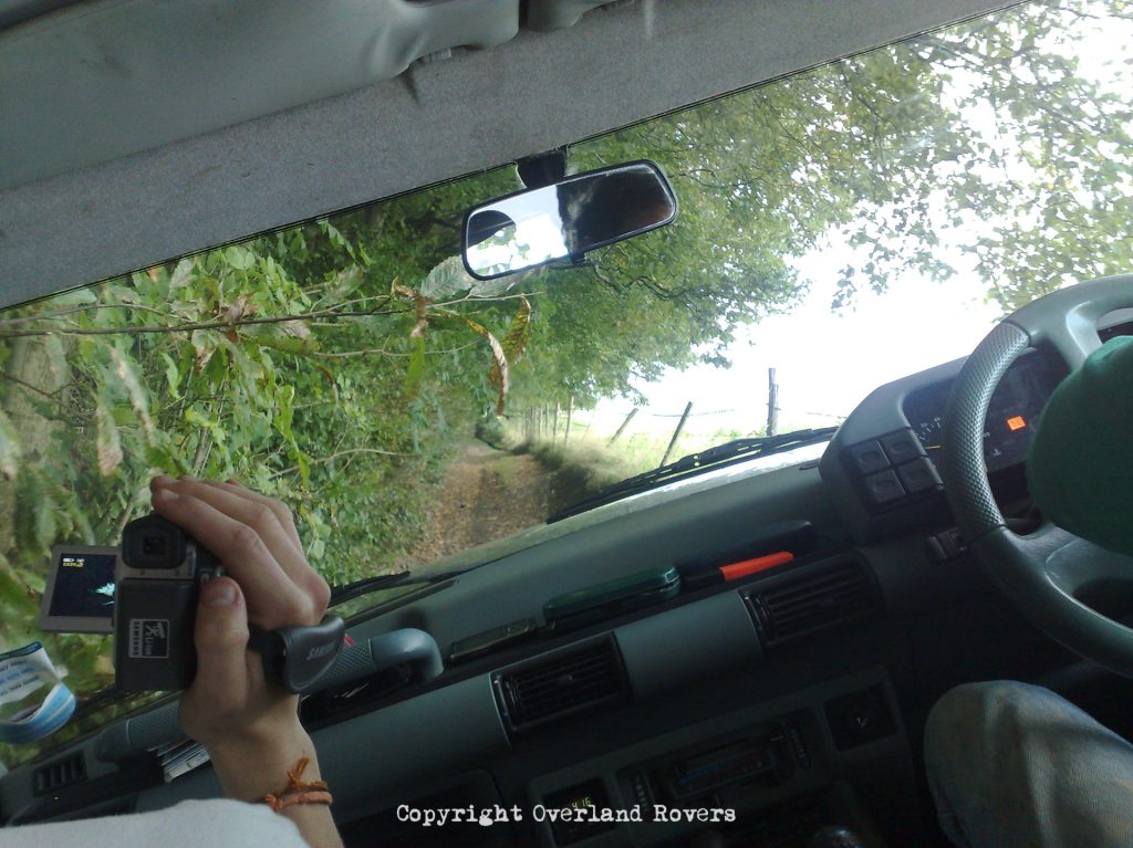Looking out the front of a Land Rover Discovery 1, from the rear seat, with the dashboard in view. There is a dirt covered trail leading into the distance, the car is leaning heavily to the left, and a hand is visible from the passenger, holding a video camera which is pointing towards the front.