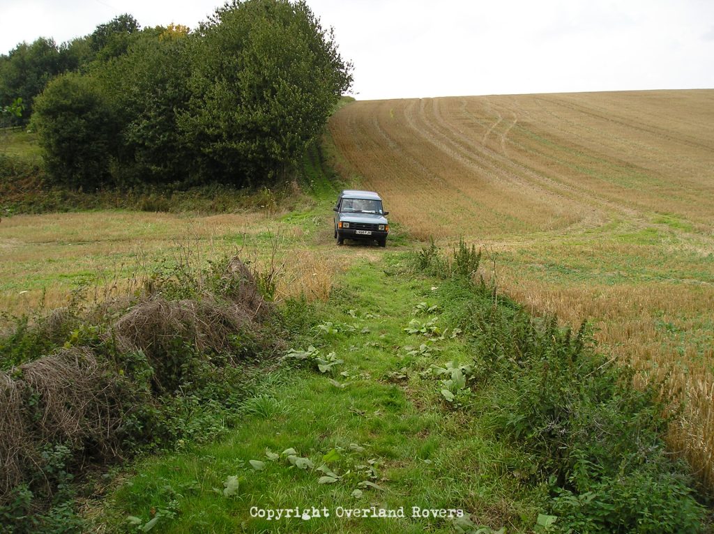 A blue Land Rover Discovery 2oo Tdi in the distance is facing the camera. The vehicle is on a grass track with trees to the left behind it, and a harvested field to the right