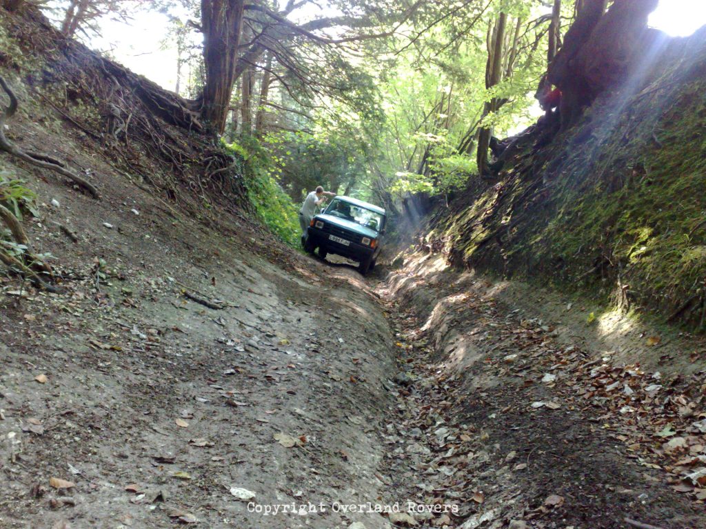 Looking down a steep sided dirt track, with trees either size, at a blue Land Rover Discovery 1 in the distance, leaning to one side
