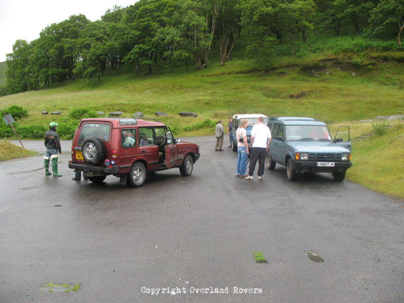 One blue, one red, Land Rover Discovery 1 parked in a car park with various people milling around.