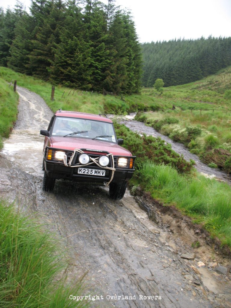 A red 1993 Land Rover Discovery climbing a rock slope, with trees in the background