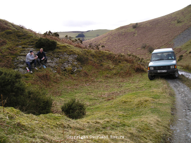 2 men sitting on a green slope eating lunch, with a blue Land Rover Discovery 1 sat on a gravel track to the right. There are green hills in the background