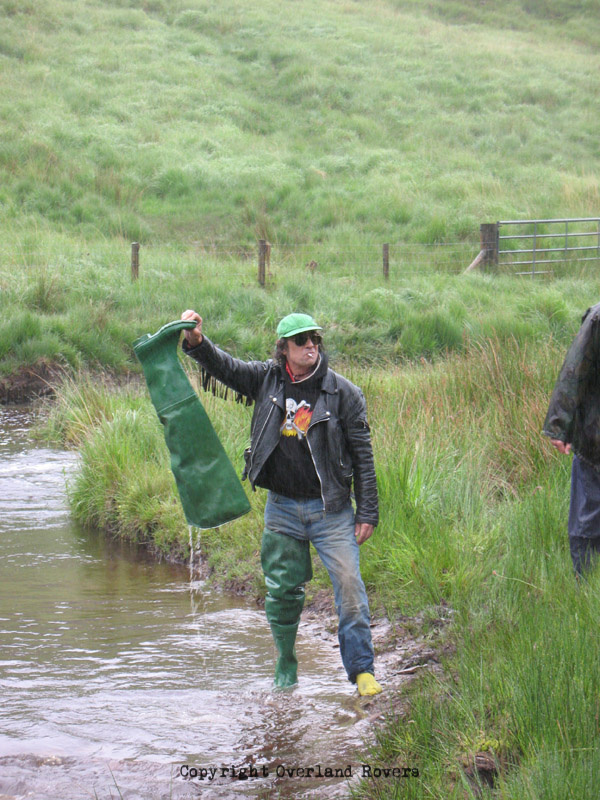 A man standing at the side of a stream, wearing one wader and emptying the other one of water.