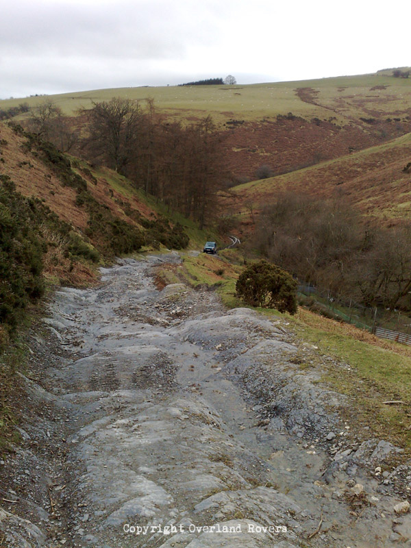 A blue Land Rover Discovery 1, in the distances, climbing a steep rocky green lane in Wales. There are rolling green hills surrounding the scene.