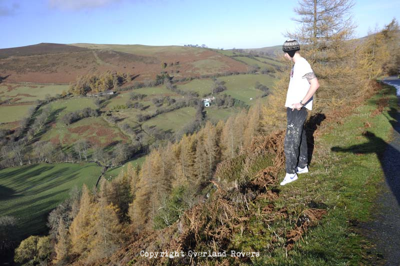 A man dressed in white t-shirt with a woolly hat and hands on hips, looks out across a Welsh valley.