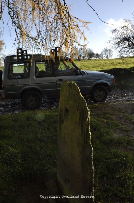 A blue Land Rover Discovery 1, 3 door, on a track, with a tree hanging overhead and a tombstone in the foreground